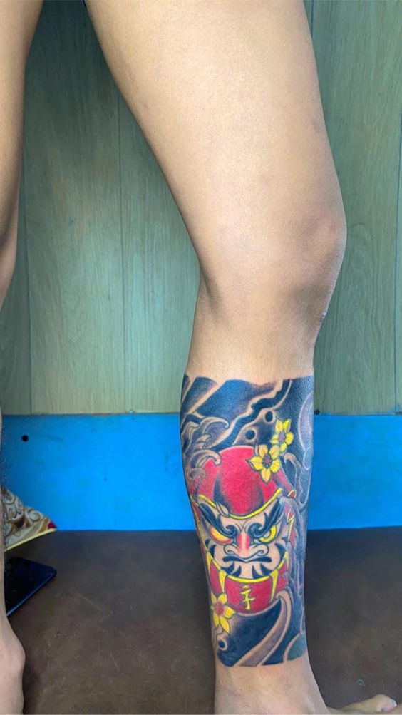 Hình xăm Daruma kết hợp màu sắc nổi bật Hình xăm mặt nạ Nhật Bản trên cẳng chân, phong cách màu sắc chi tiết, độ phân giải HD.