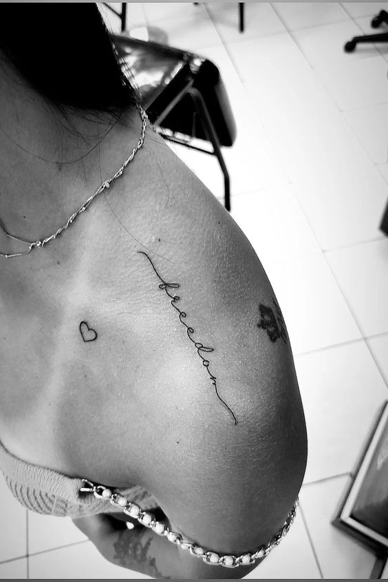 Hình xăm chữ "freedom" phong cách minimalist trên vai, cùng một hình trái tim nhỏ, ảnh đen trắng HD.
