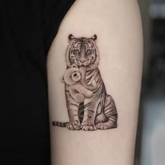 Hình xăm hổ ôm gấu bông trên cánh tay, phong cách blackwork chi tiết. Ảnh chất lượng HD.