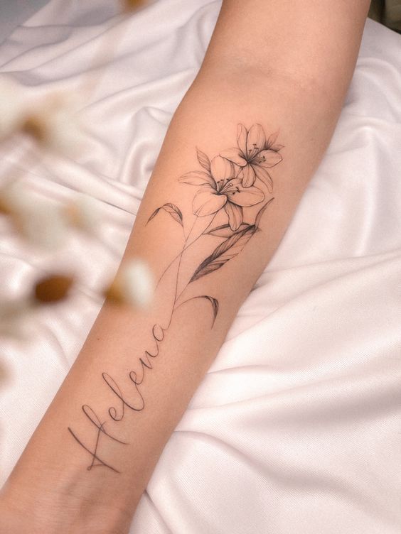 Hình xăm hoa ly mini kèm tên tạo sự độc đáo Hình xăm hoa trên cánh tay, phong cách blackwork tinh tế, chi tiết rõ nét, ảnh độ phân giải cao.