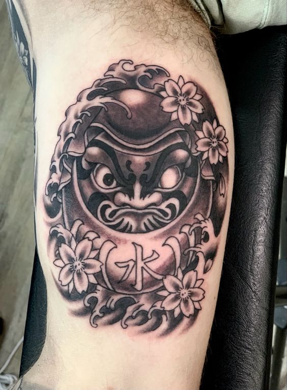 Hình xăm daruma một mắt kết hợp với hoa đào, mang tính nghệ thuật cao Hình xăm mặt nạ Daruma trên bắp tay, phong cách blackwork, chi tiết sắc nét, độ phân giải HD.