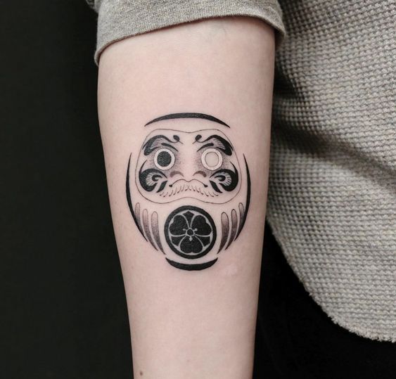 Mẫu xăm dành cho phái nữ, hình daruma gắn cỏ ba lá, mang lại sự thuận lợi Hình xăm Daruma trên cánh tay, phong cách blackwork, chi tiết rõ nét, độ phân giải HD.