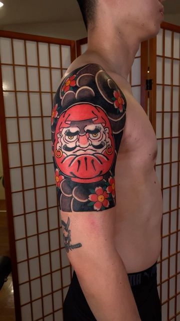 tác phẩm xăm daruma với nét mặt giận dữ hòa quyện cùng hoa anh đào đỏ đậm chất Nhật Bản Hình xăm Daruma phong cách Nhật Bản, vị trí bắp tay, màu sắc tươi sáng, độ chi tiết cao, ảnh HD.